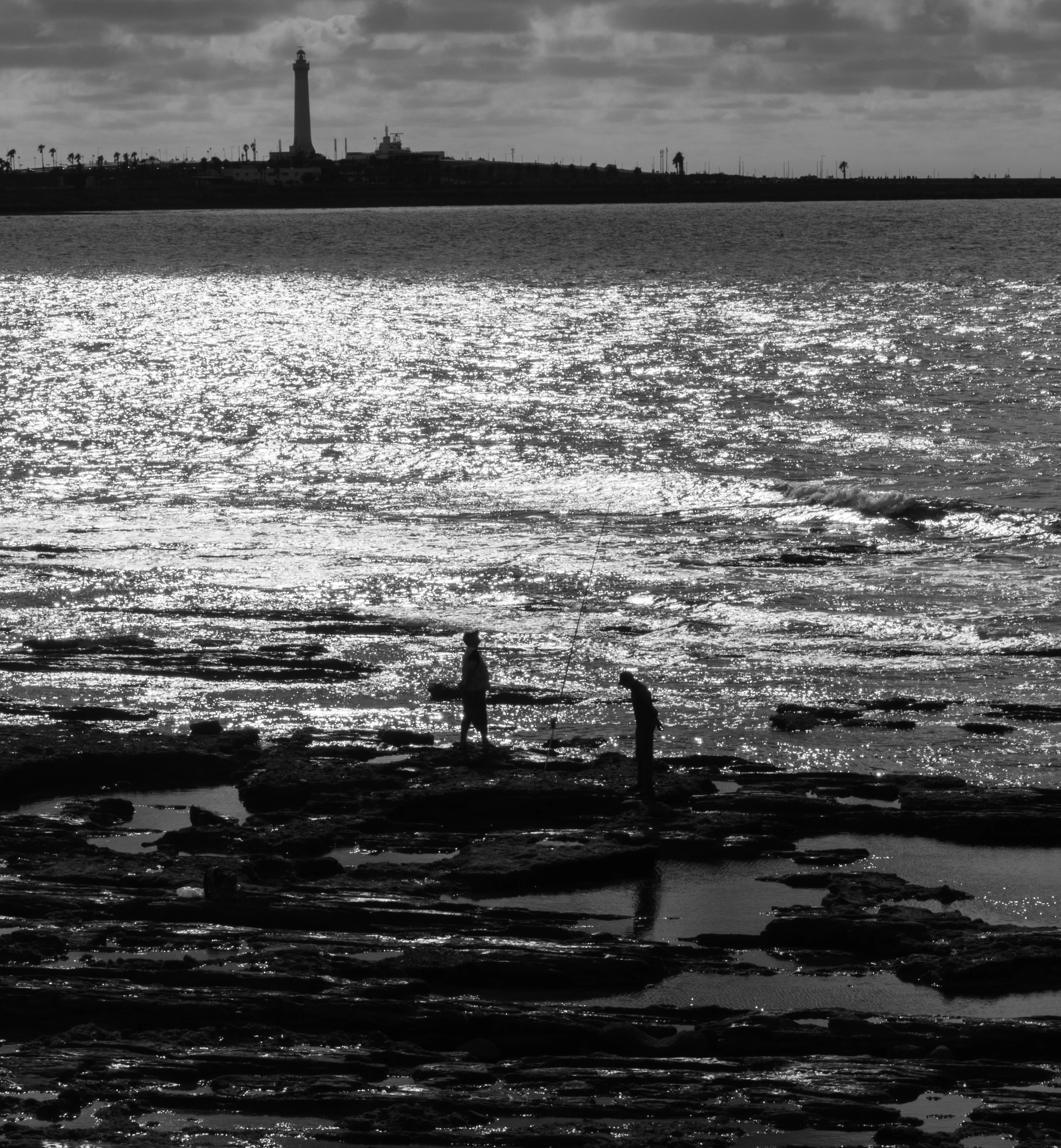 Two silhouetted figures fish from tidal rocks with a lighthouse across the water