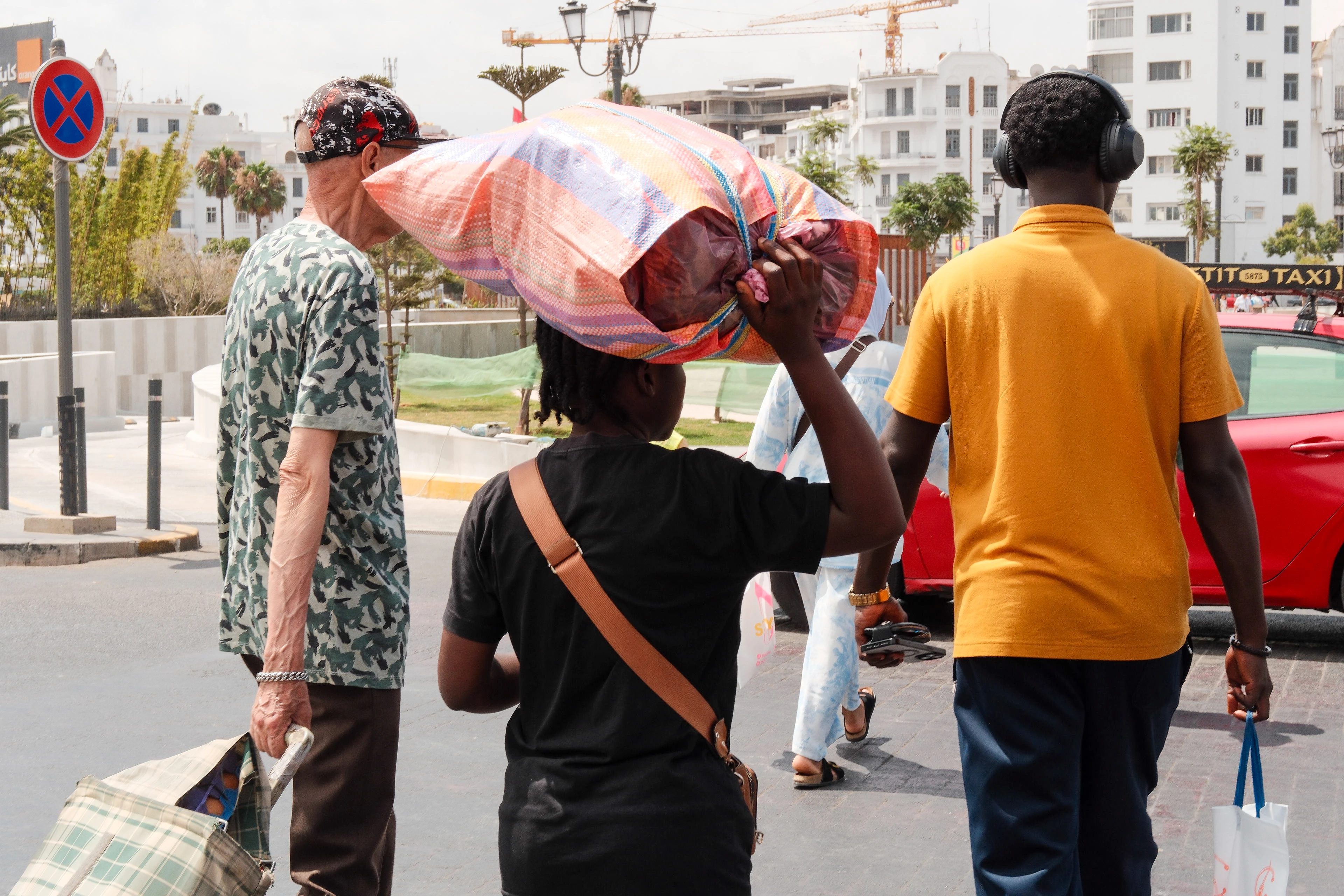 Pedestrians cross a bright street carrying bags and a large wrapped bundle