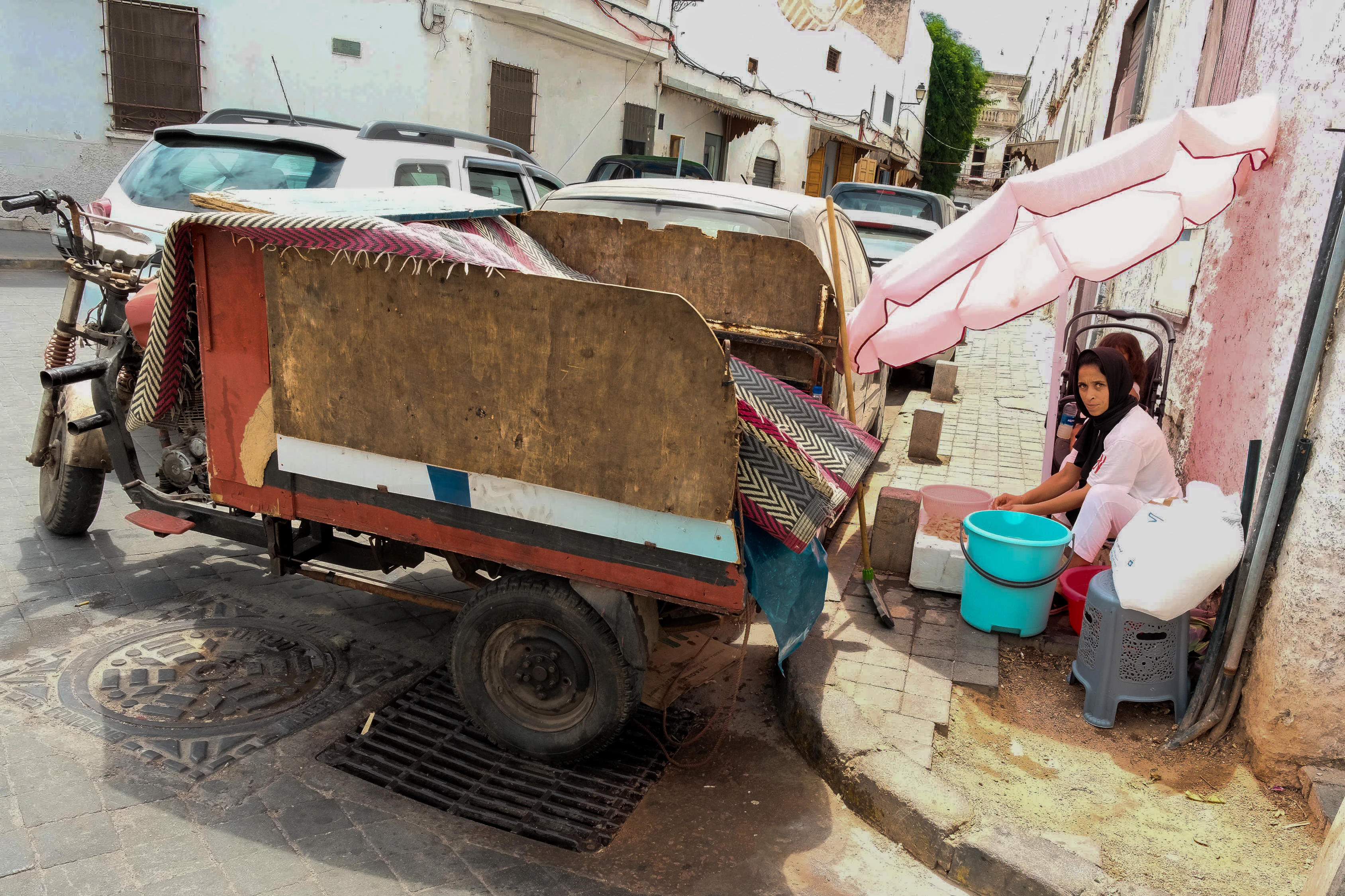 A loaded three-wheeled cart stands beside a seated woman under a pale umbrella