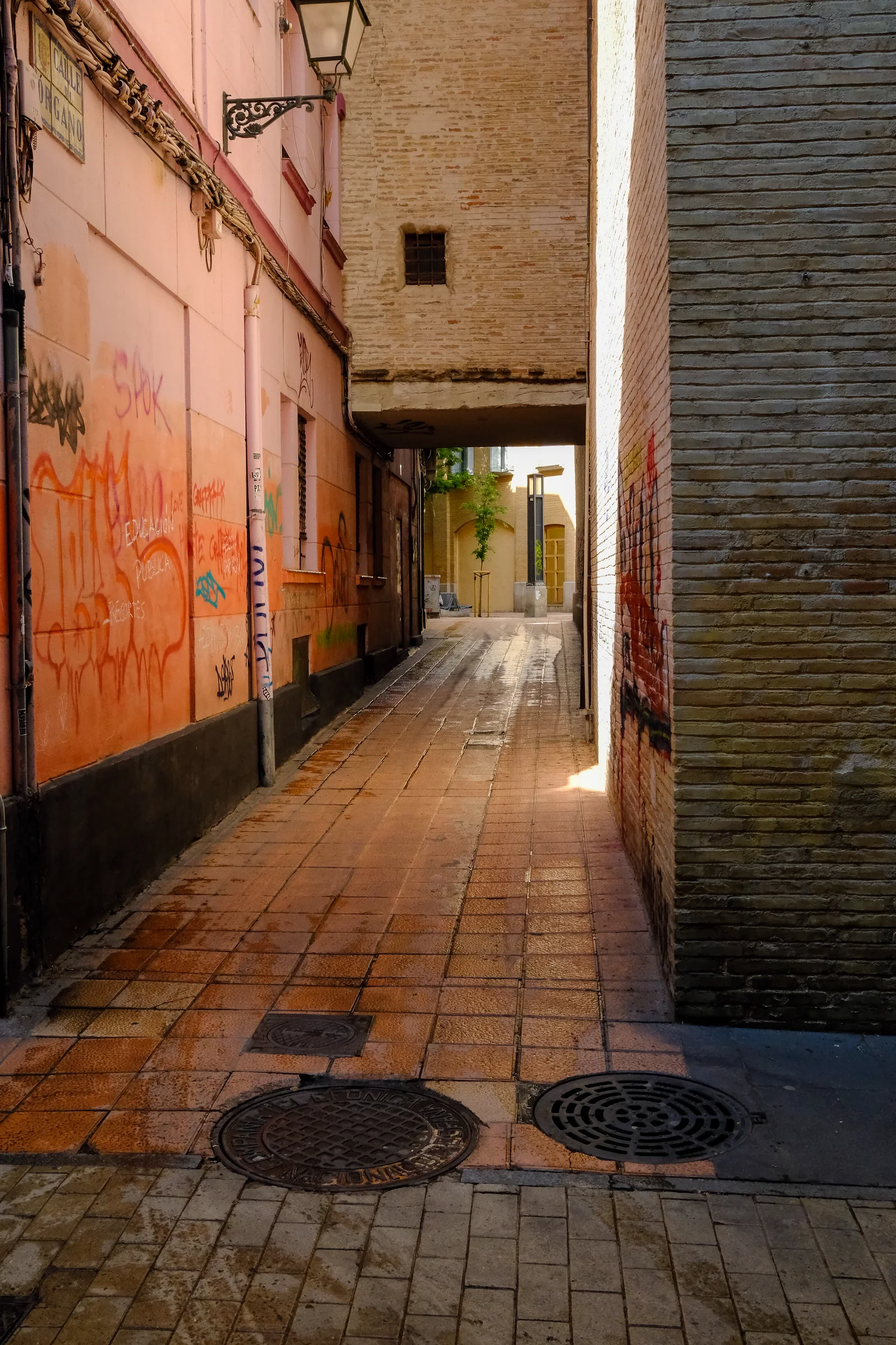 A narrow wet passage between warm graffiti-marked walls and brick