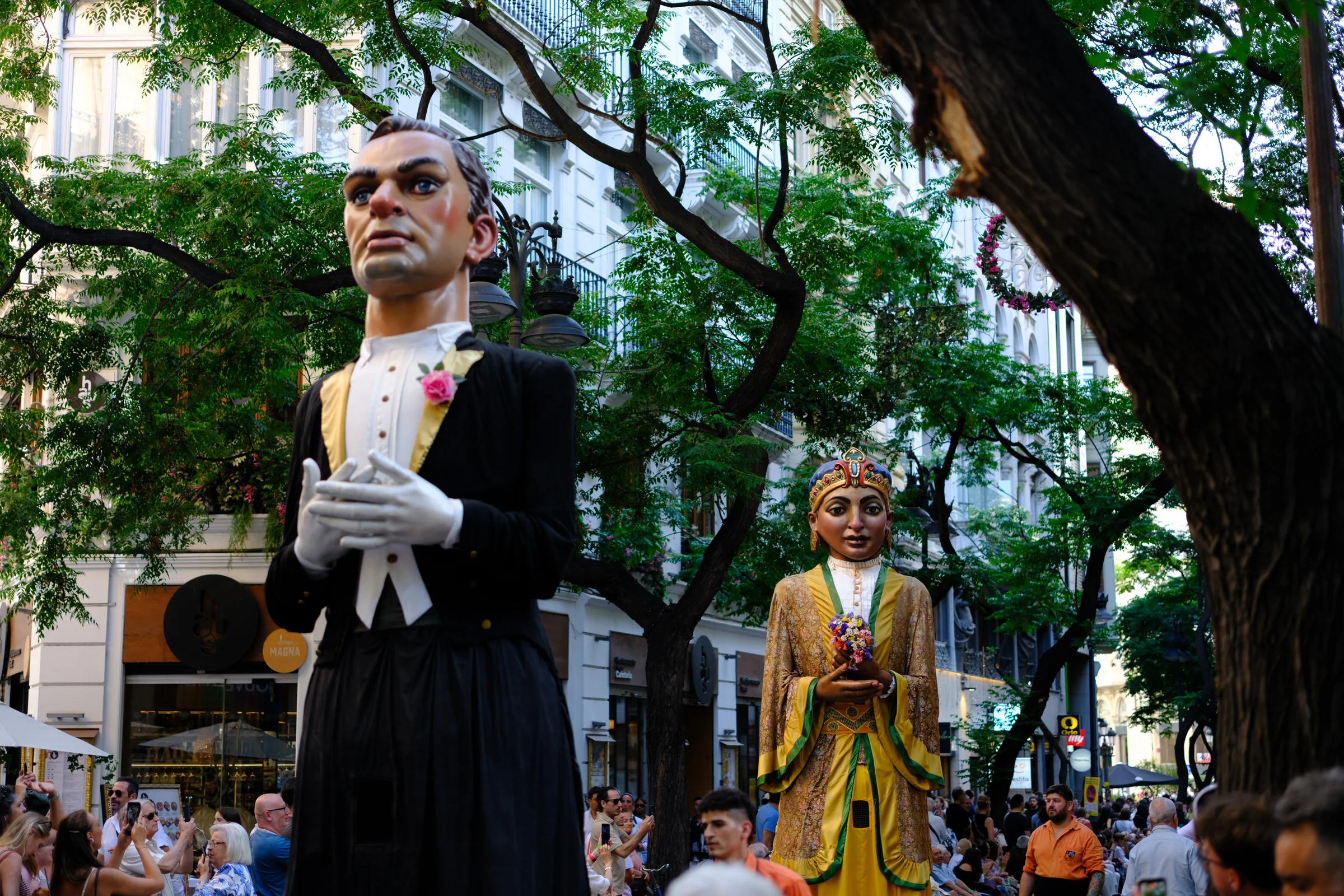 Two giant festival figures stand among trees and a dense street crowd