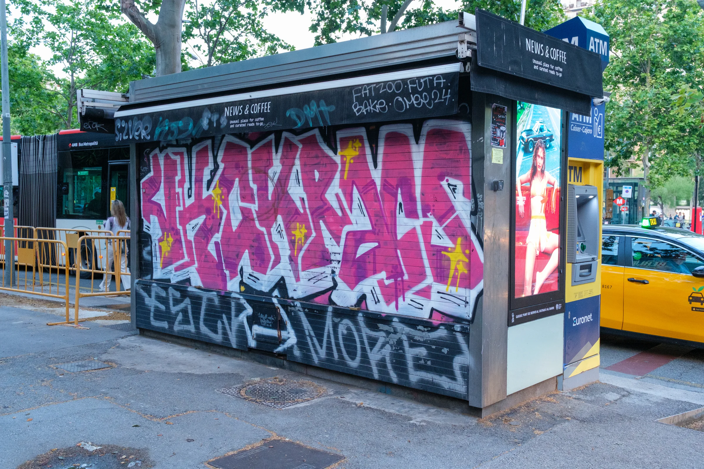 A closed news kiosk covered with pink graffiti beside an ATM and yellow taxi