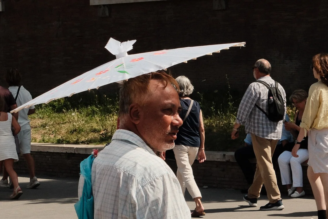 A man in profile walks under a white parasol in hard sunlight