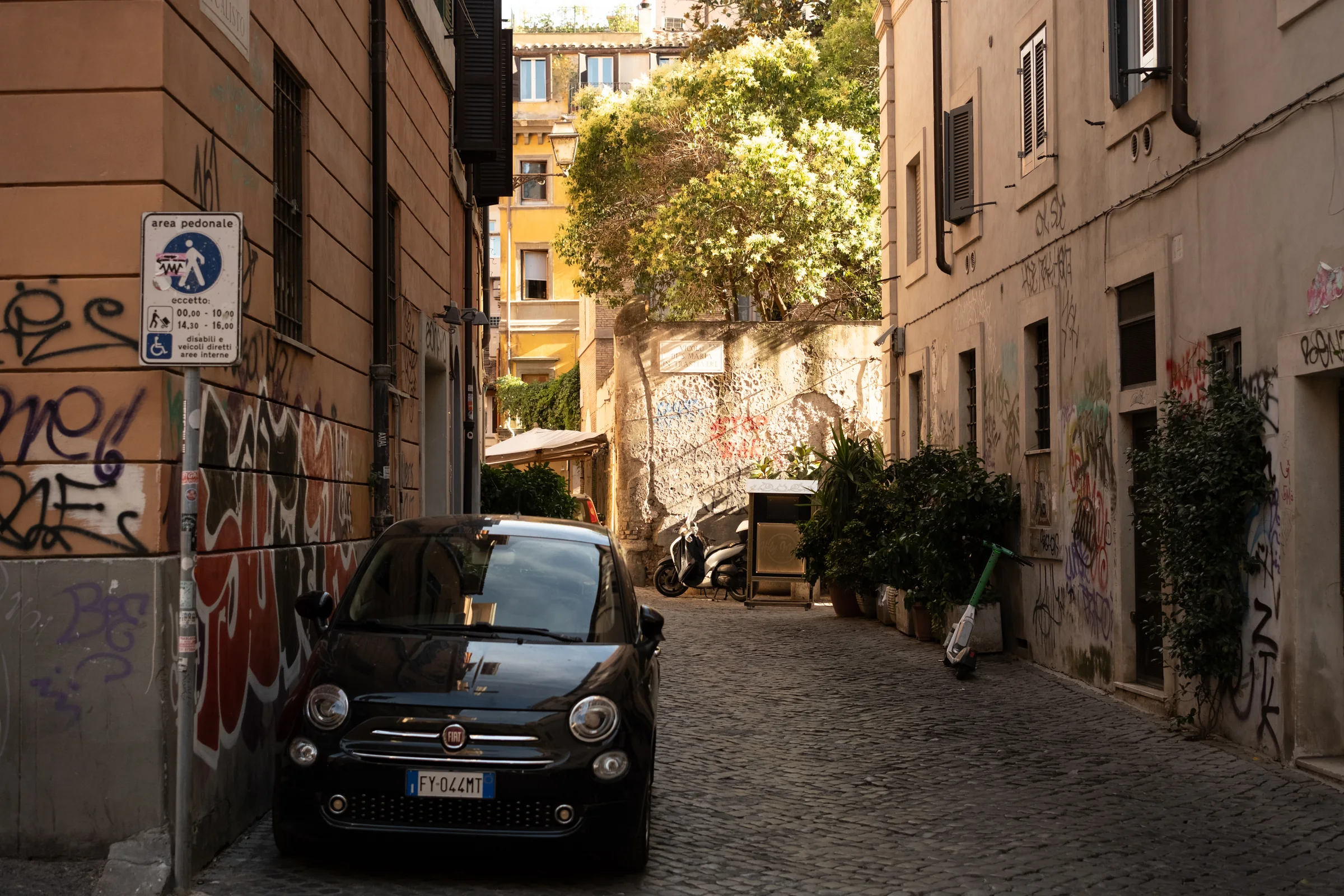 A compact car and scooter sit in a warm narrow Roman street covered with graffiti